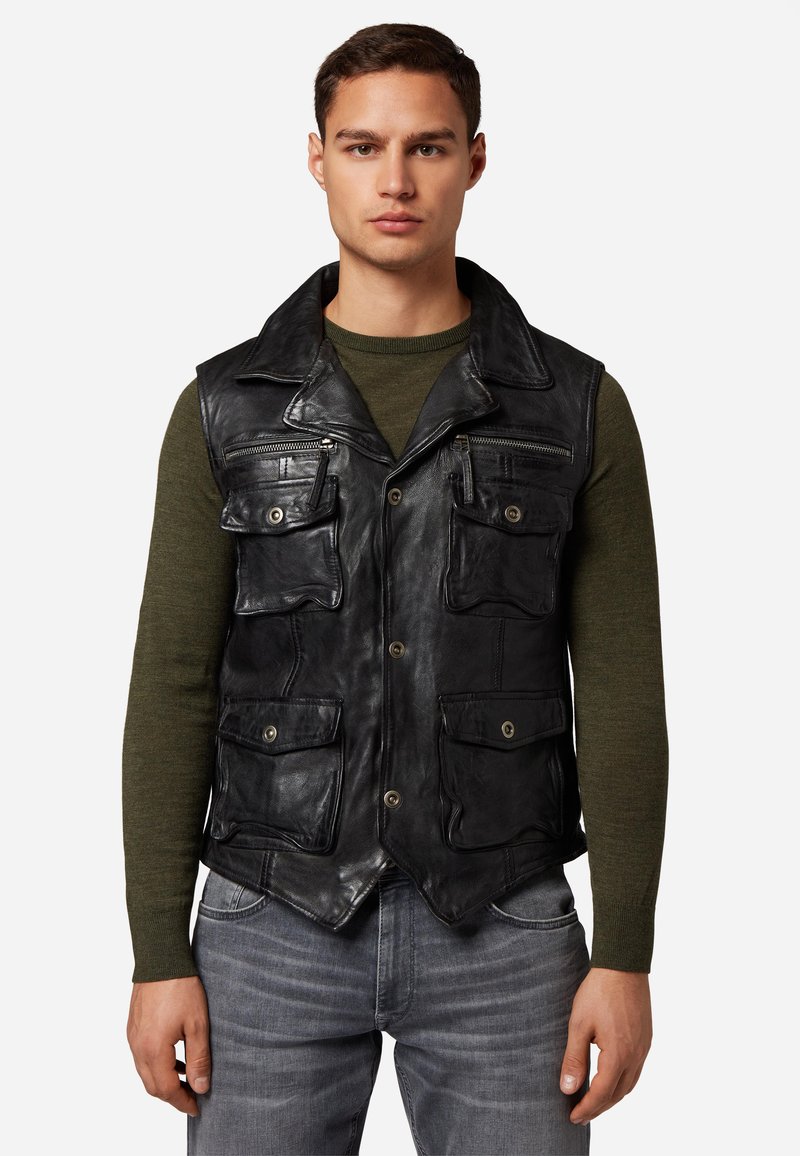 Schwarze Lederweste mit mehreren Fronttaschen und einem Kragen. Getragen über einem grünen Langarmshirt mit strukturierter Oberfläche.