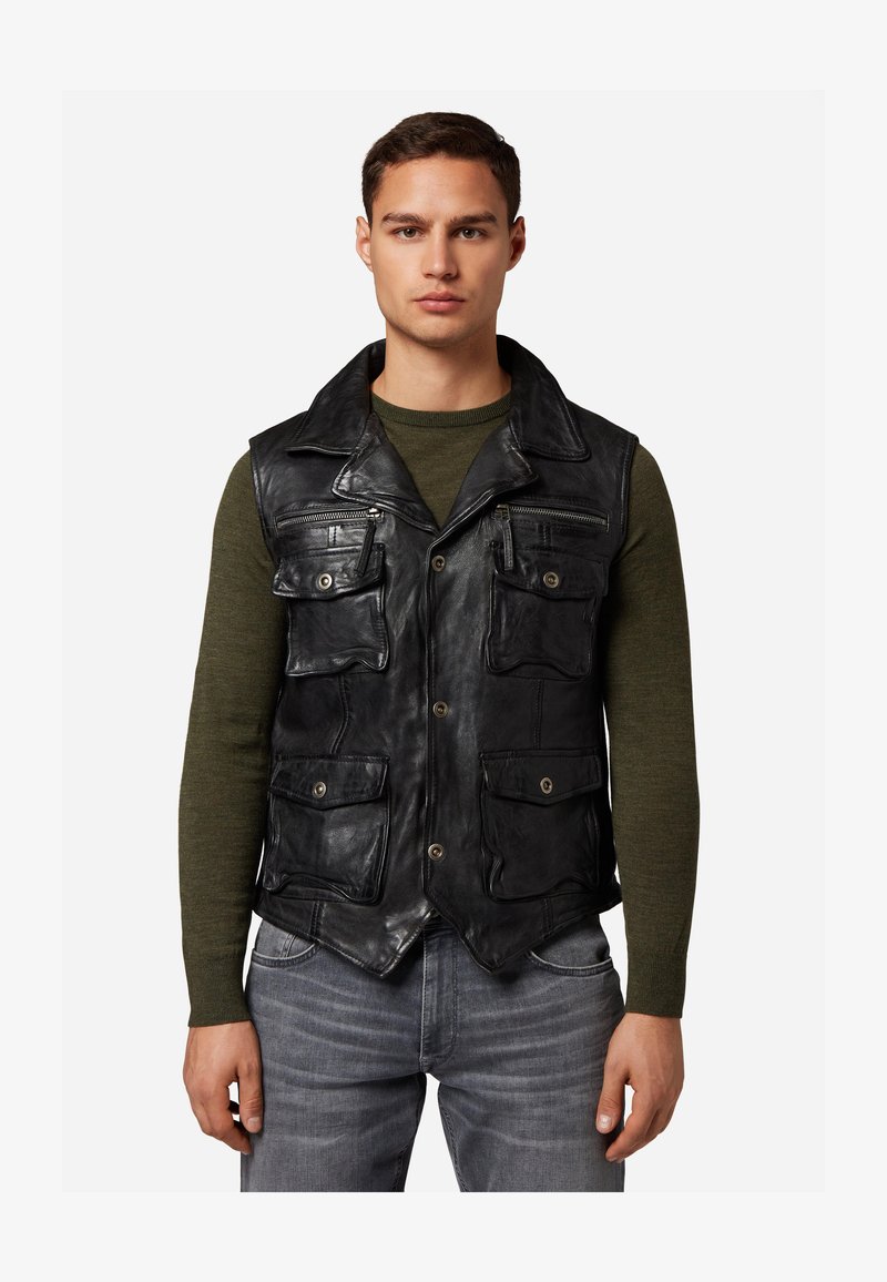 Schwarze Lederweste mit mehreren Fronttaschen und einem Kragen. Getragen über einem grünen Langarmshirt mit strukturierter Oberfläche.