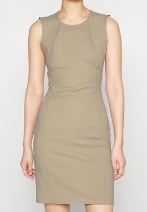 Vestido beige sin mangas hasta la rodilla con escote redondo y cintura ajustada, llevado por una persona que está de pie frente a un fondo liso.