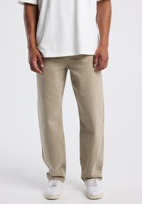 Hombre vistiendo pantalones rectos beige, zapatillas deportivas blancas y una camiseta blanca, de pie frente a un fondo claro y liso.