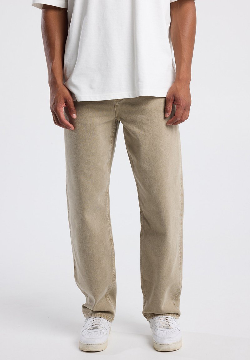 Hombre vistiendo pantalones rectos beige, zapatillas deportivas blancas y una camiseta blanca, de pie frente a un fondo claro y liso.