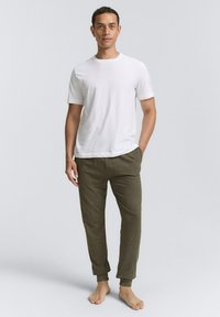 Witte t-shirt met korte mouwen en ronde hals, gecombineerd met olijfgroene tapered sweatpants met elastische boorden. Model staat op blote voeten.