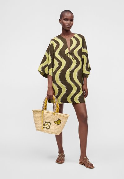 Femme portant une robe ample à motif ondulé vert et jaune, tenant un sac en paille tressée avec des motifs de bananes et de patchs, et portant des sandales marron.
