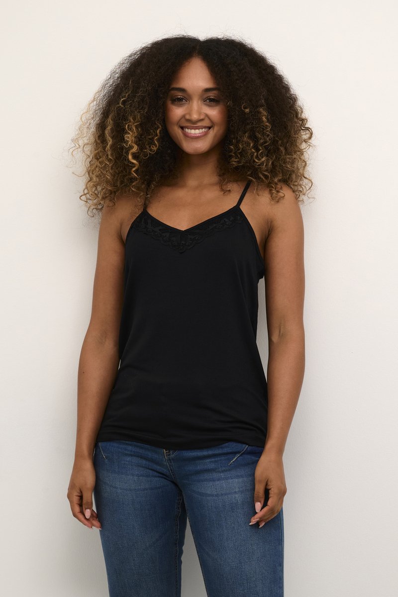 Femme aux cheveux bouclés souriante, portant un haut noir sans manches avec bordure en dentelle et un jean bleu, debout sur un fond blanc.