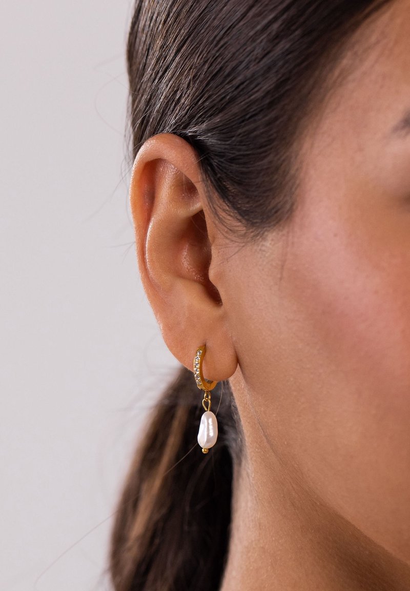 Gros plan sur l'oreille d'une femme aux cheveux foncés portant une boucle d'oreille créole en or ornée de petits cristaux et d'une perle blanche pendante.
