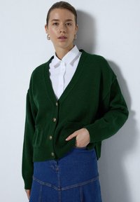 Groene gebreide cardigan met een V-hals en knoopsluiting, is voorzien van twee voorzakken, geribde manchetten en een ontspannen pasvorm. Witte blouse eronder.