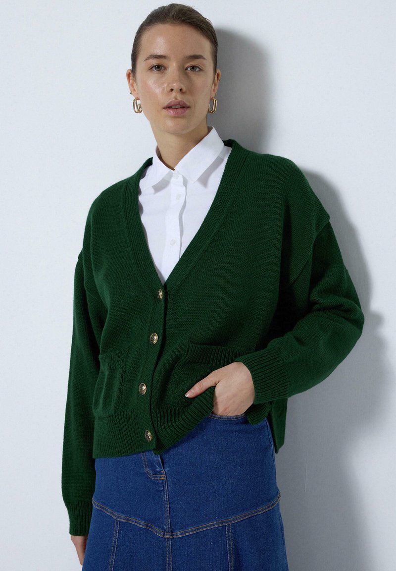 Zelený pletený cardigan s výstřihem do V a zapínáním na knoflíky, má dvě přední kapsy, žebrované manžety a volný střih. Pod ním je bílá blůza.