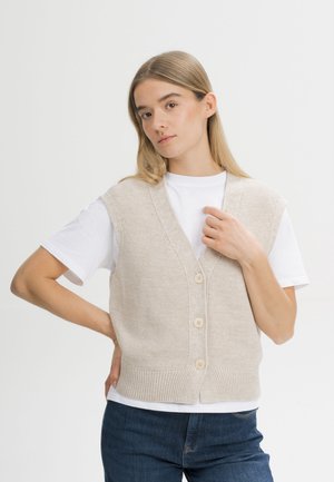 Jonge vrouw met blond haar draagt een beige gebreid vest met knopen over een wit T-shirt en blauwe spijkerbroek tegen een witte achtergrond.