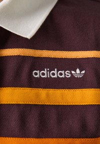 Bordo polo majica s bijelim ovratnikom, s horizontalnim narančastim i žutim prugama. Izvezeni "adidas" logotip u bijeloj boji na prsima.
