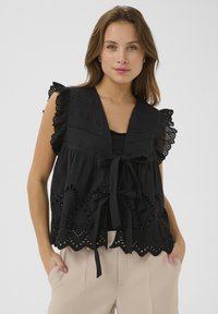 Blusa nera in cotone con maniche decorate da pizzo, scollatura a cravatta e motivi a occhio decorativi lungo l'orlo. Abbinata a pantaloni beige.