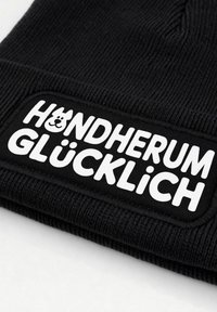 Black knit beanie with a textured fabric. Features a white patch with the text "HUNDERUM GLÜCKLICH" and a bear graphic.