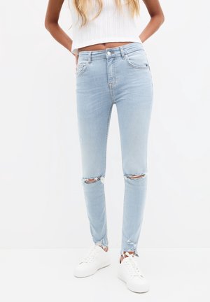 Femme portant un jean skinny déchiré bleu clair, un top court en maille blanche et des baskets blanches, debout sur un fond blanc.