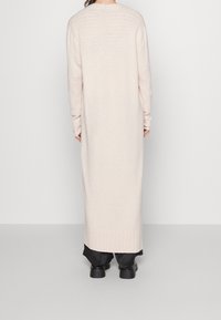 Długi, jasnoróżowy kardigan z prążkowanymi mankietami i dołem. Miękka faktura, pełna długość i lekko luźny krój, noszony na ciemnych spodniach.