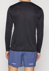 Mies, który ma na sobie czarną długą koszulkę sportową i niebieskie krótkie spodenki do biegania, widoczny z tyłu na jednolitym tle.
