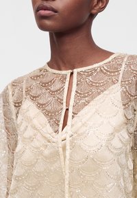 Gros plan d'une personne portant un haut transparent beige brodé de sequins, avec un petit décolleté goutte d'eau et un détail de fermoir.
