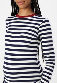 Langermet topp med marineblå og hvite horisontale striper, med rød rund hals. Myk stoff, figursydd design, egnet for ulike anledninger.