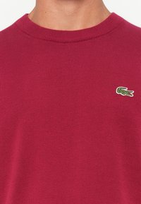 Pulôver de malha bordeaux com gola redonda canelada, textura macia e um pequeno logótipo de crocodilo bordado em verde no peito.
