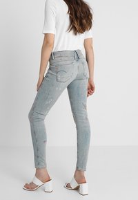 Bleka ljusgrå skinny jeans med slitna detaljer och färgglada färgfläckar, matchat med vita klacksandaler och en kortärmad topp.