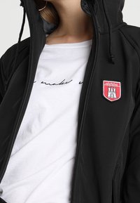 Giacca nera con cappuccio, chiusura con zip e cordini, caratterizzata da un patch ricamato rosso. Indossata sopra una camicia bianca con del testo.