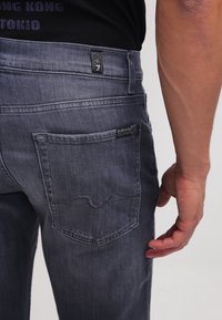 Jeans de denim gris oscuro con un diseño ajustado, que cuentan con un bolsillo trasero con detalles de costura y una etiqueta de la marca cerca de la cinturilla.