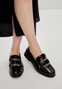 Mocassins en cuir verni noir avec deux accents de chaînes métalliques sur le devant, texture lisse, bout arrondi et semelle en caoutchouc à profil bas.