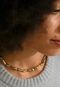 Collana dorata con un design irregolare e texture, caratterizzata da elementi collegati a forma organica. Indossata sopra un maglione grigio.