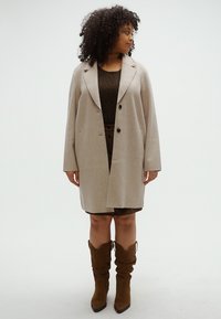 Μπεζ υπερμεγέθες παλτό με γιακά με σχισμές, δύο κουμπιά και πλαϊνές τσέπες, φορεμένο πάνω από ένα καφέ τοπ με σχέδια και καφέ φούστα, συνδυασμένο με καφέ χαλαρά μποτάκια.