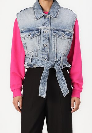 Person trägt ärmellose, verwaschene Jeansjacke mit Knöpfen vorne und gebundenem Gürtel über einem knallpinken Langarmshirt und schwarzen Hosen.