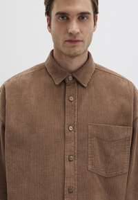 Braunes Cordhemd mit Knopfleiste, spitzem Kragen und einer einzelnen Brusttasche. Vertikale gerippte Textur und Holzknöpfe.