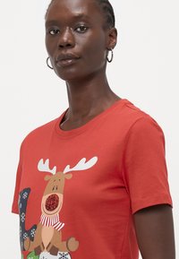 Raudona medvilninė marškinė su animaciniu elnio grafiku, turinčiu ragus, dryžuotą šalį ir snaiges. Trumpi rankoviai ir apvalus iškirpimas.