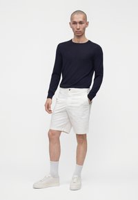Homme debout, une main dans la poche, portant une chemise noire à manches longues, un short blanc, des chaussettes blanches et des baskets blanches sur fond blanc.
