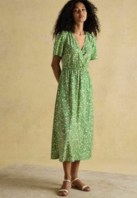 Grünes florales Midi-Kleid mit kurzen Ärmeln, elastischem Bund und V-Ausschnitt. Glattes Material mit leichter Textur; kombiniert mit neutralen Sandalen.