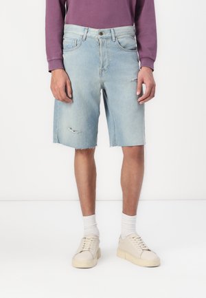 Mann trägt hellblaue, zerrissene Jeansshorts, weiße Socken, beige Turnschuhe und ein lila Langarmshirt, steht vor weißem Hintergrund.