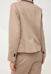 Blazer beige sur mesure avec épaules structurées, revers crantés et accents de boutons noirs sur les manches. Tissu lisse avec une texture subtile.