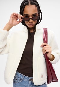 Giacca di pile bianca con colletto alto, bordi beige in contrasto, maniche oversize e tasche laterali. Il modello tiene una borsa lucida color bordeaux.