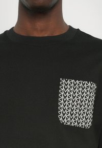 Svart T-skjorte med rund hals og venstre brystlomme med et geometrisk hvitt mønster. Stoffet ser glatt ut, av bomullsmiks.