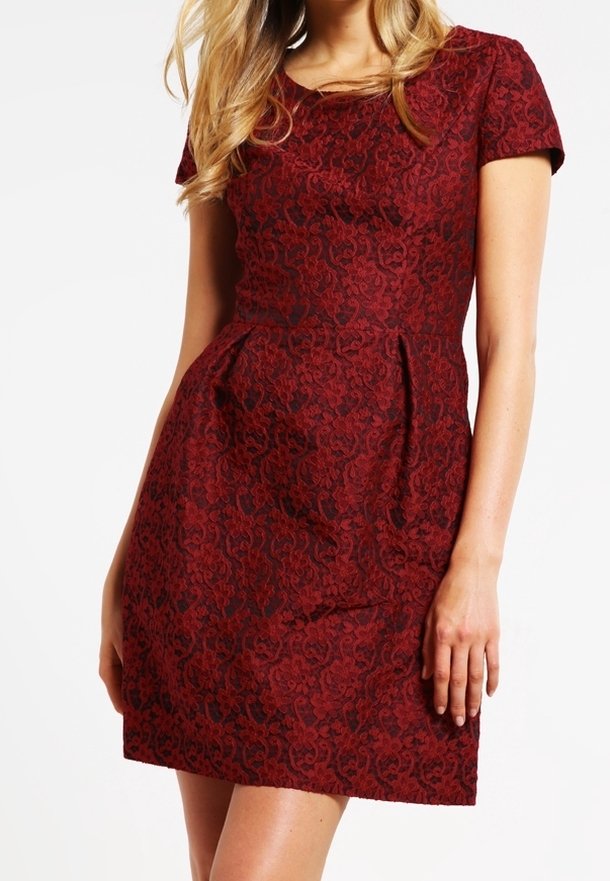Femme portant une robe rouge ajustée, à manches courtes, longueur genou, avec un motif floral subtil et des plis à la taille.