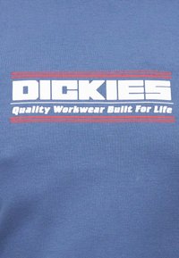 Λογότυπο Dickies με το σύνθημα «Ποιοτική Εργατική Ενδυμασία Κατασκευασμένη για τη Ζωή», κείμενο σε λευκό πάνω σε μπλε ύφασμα με κόκκινες οριζόντιες γραμμές πάνω και κάτω.