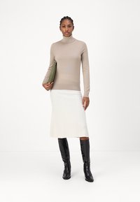 Beige garbónyakú pulóver hosszú ujjakkal, krém színű bordázott midi szoknyával párosítva. A modellt egy zöld clutch táska és fekete combcsizma díszíti.