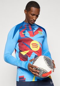 Niebieska, długorękawowa koszulka sportowa z kolorowym abstrakcyjnym wzorem, w tym geometryczne kształty w kolorze czerwonym, żółtym i niebieskim. Model trzyma wzorzystą piłkę nożną.