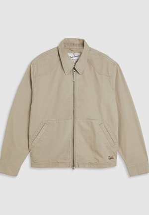 Chaqueta de lona beige con cremallera frontal, cuello puntiagudo y dos bolsillos laterales. Presenta detalles de costura y un pequeño logo en el dobladillo.