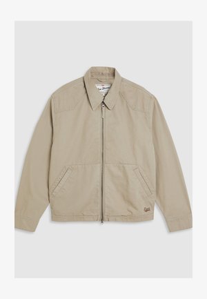 Chaqueta de lona beige con cremallera frontal, cuello puntiagudo y dos bolsillos laterales. Presenta detalles de costura y un pequeño logo en el dobladillo.