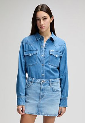 Fiatal nő hosszú sötét hajjal, gombos kék farmeringben és világoskék farmer szoknyában, egyszerű háttér előtt.