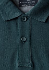 Tmavě modré polo tričko s texturovanou tkaninou, žebrovaným límcem, dvěma černými knoflíky a logem na vnitřní části límce.