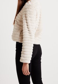 Krämfärgad faux fur-jacka med en texturerad, ribbad design, cropped längd och långa ärmar. Kombination med svarta, åtsittande byxor.