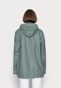 Henkilö, jolla on lyhyet ruskeat hiukset ja päällä vaaleanvihreä huppari sadetakki ja valkoiset housut, seisoo vaaleanharmaata taustaa vasten.