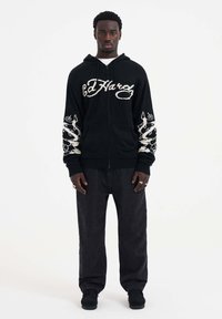 Hoodie zippé noir avec logo Ed Hardy blanc et motifs complexes sur les manches ; associé à un pantalon noir ample et des chaussures noires.