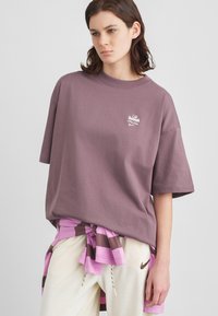 Mlad človek nosi prevelik mauve T-shirt s podpisom "Café Swoosh", črtasto majico zavezano okoli pasu in smetanove trening hlače z logotipom Nike.
