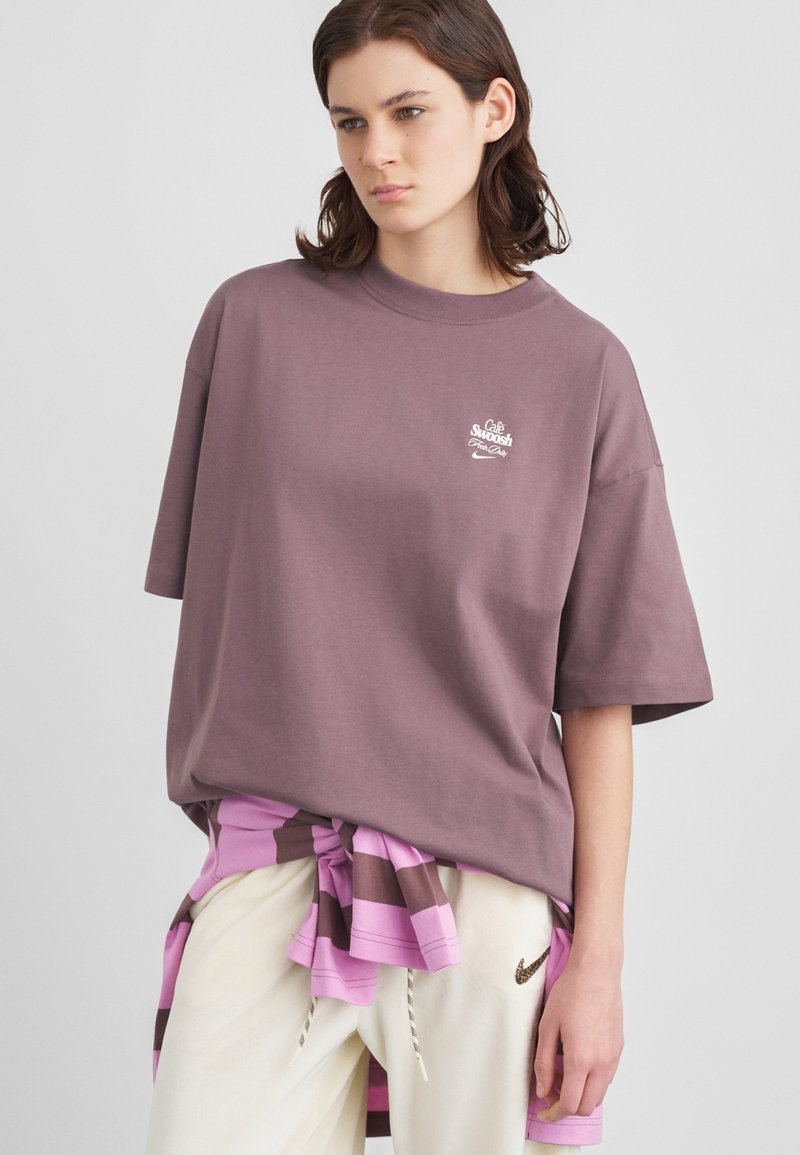 Mlad človek nosi prevelik mauve T-shirt s podpisom "Café Swoosh", črtasto majico zavezano okoli pasu in smetanove trening hlače z logotipom Nike.
