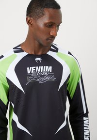 Un homme portant un t-shirt de course Venum noir, blanc et vert avec logo, regardant vers le bas sur un fond clair uniforme.